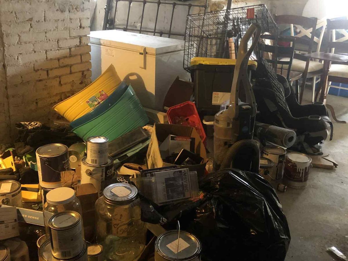 An old brick basement crowded with a chest freezer, storage bins, a vacuum, a car seat, dining chairs and stacked cans