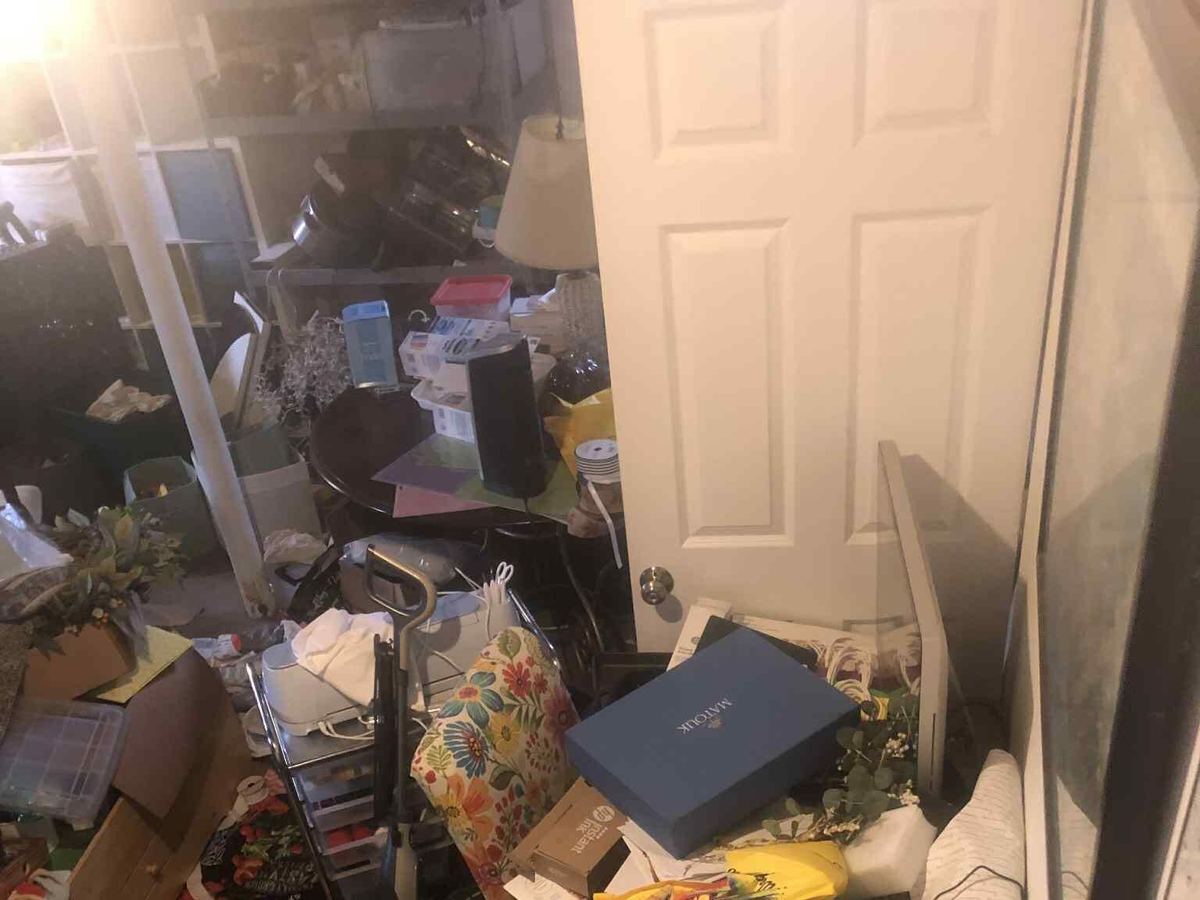 A spare room piled with boxes, bags, a cart, a lamp and household items up to the door